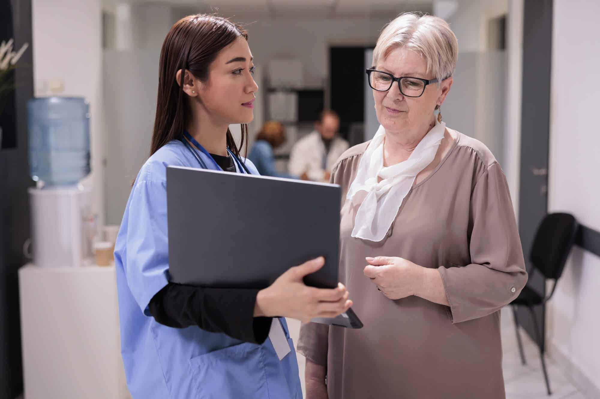 Nurse Case Manager talking about disease with old patient in hospital reception lobby, showing diagnosis on laptop to give recovery treatment.