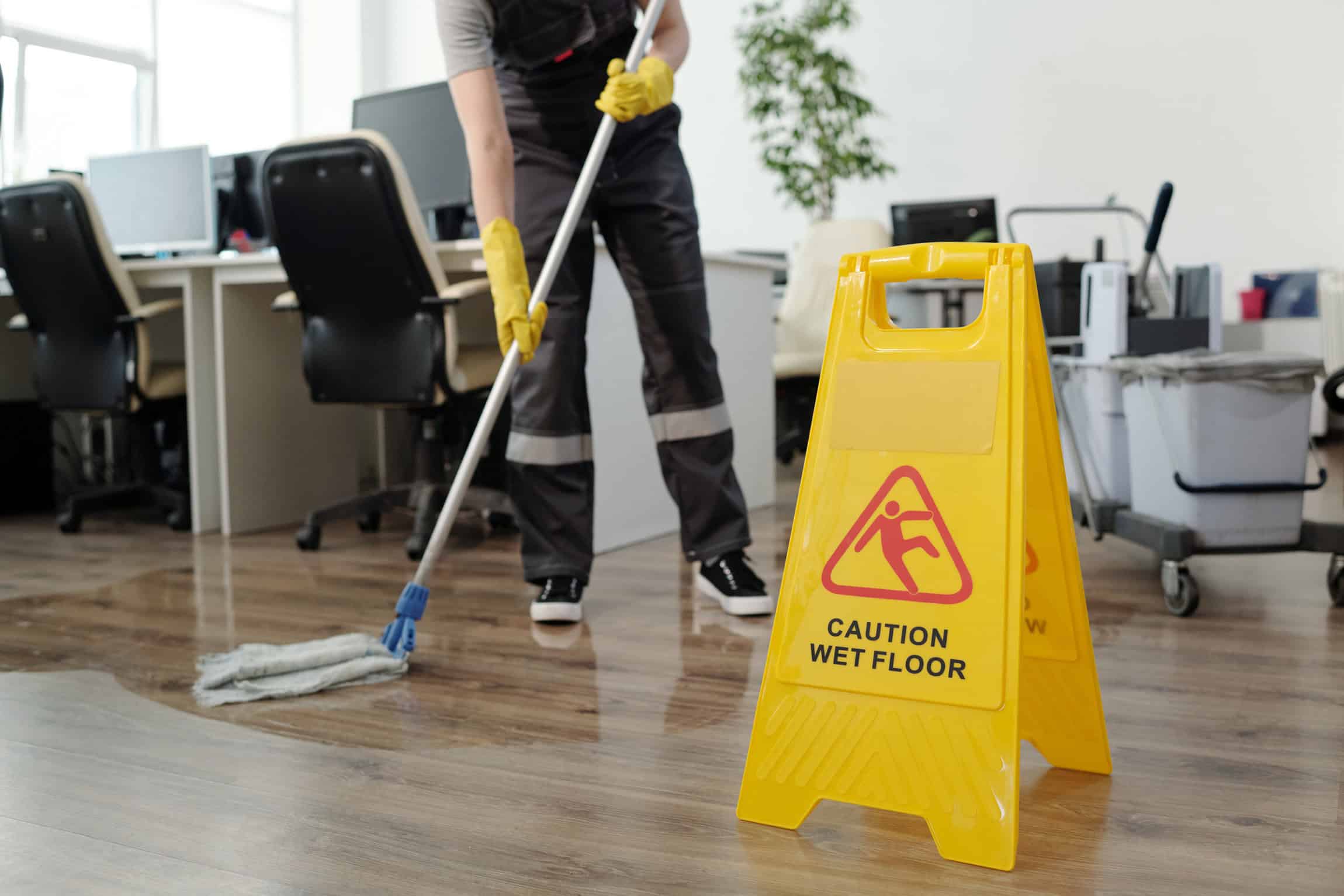 Yellow plastic signboard with caution warning about wet floor and female cleaner in black uniform working with mop in openspace office